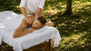 A person receiving a massage outdoors on a wooden platform with a white sheet, surrounded by trees.