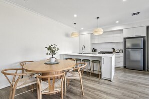 A modern kitchen with white cabinetry, a stainless steel refrigerator, and a round wooden dining table with six chairs. - Coastal Haven at Charlemont (Charlemont)