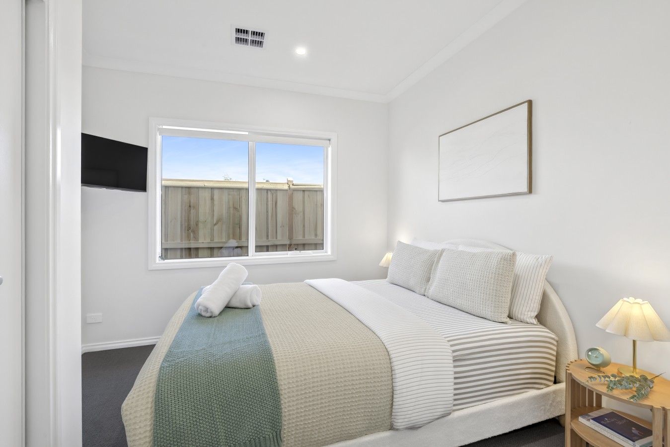 A bedroom with a bed, a bedside table with a lamp, a window with a view, and a framed picture on the wall.