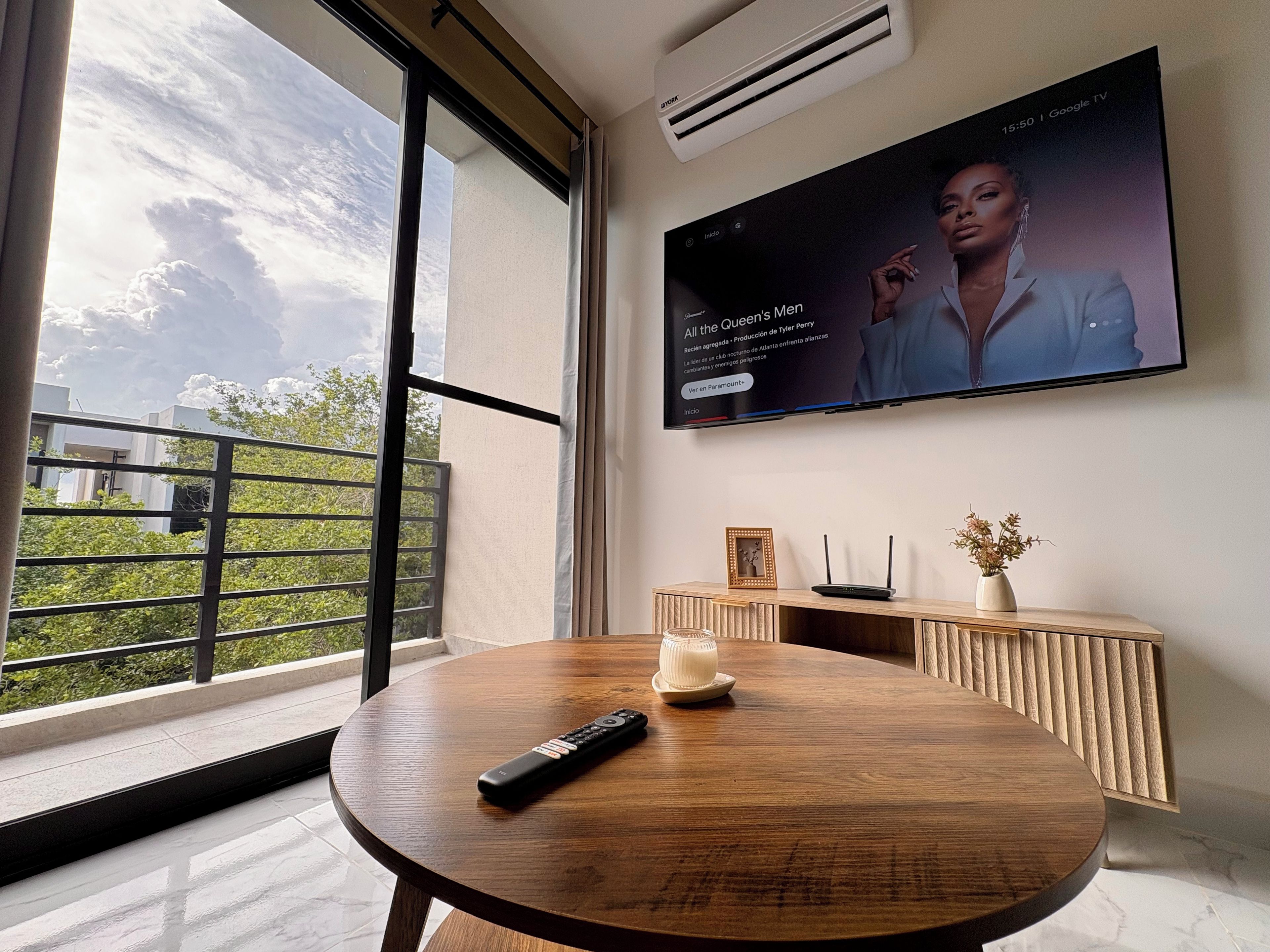 A modern living room with a large TV mounted on the wall, a round wooden table with a remote control, a decorative vase, and a framed picture on a wooden console.