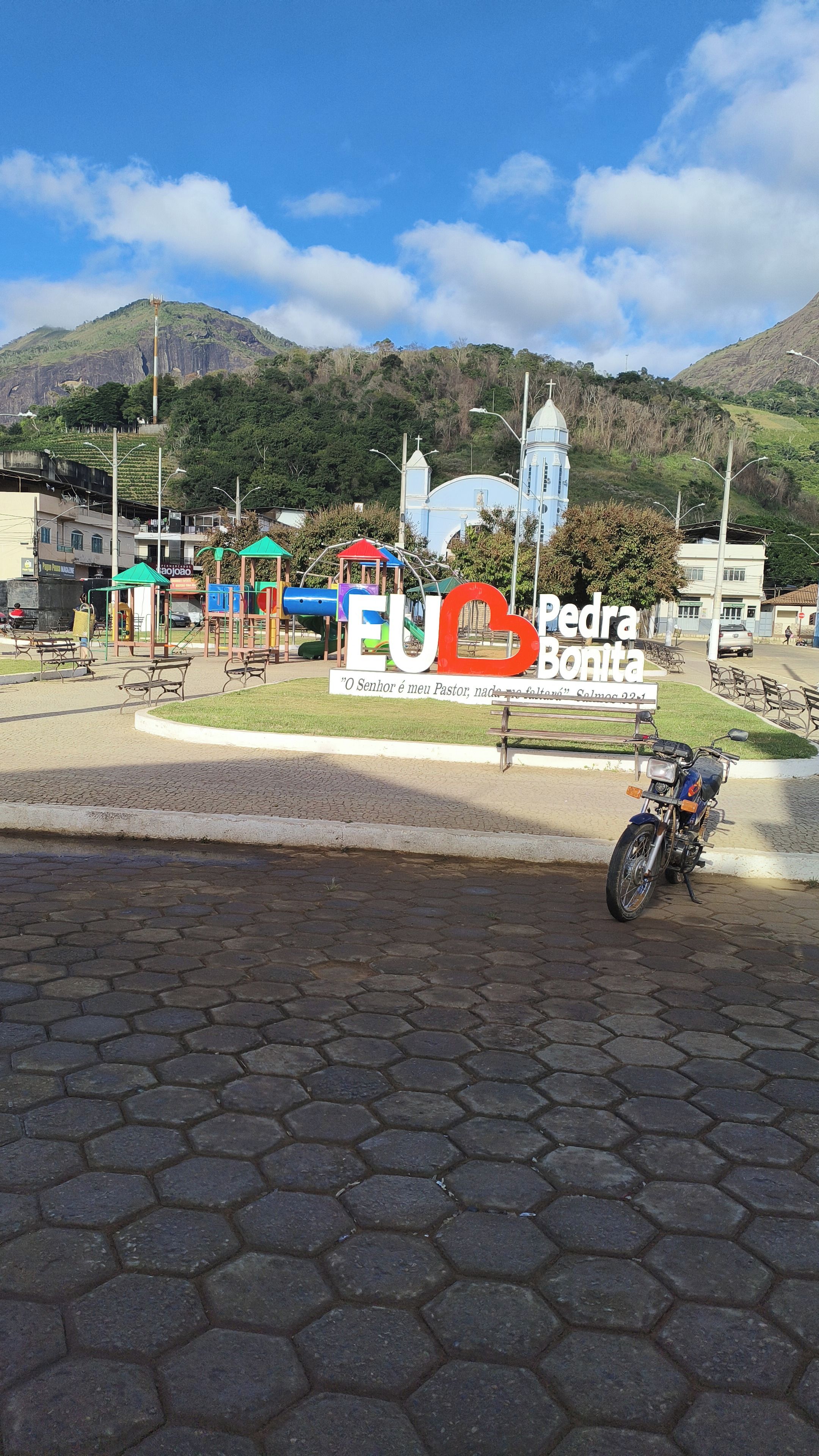 Área pavimentada com azulejos hexagonais, placa com 'EU Pedra Bonita' e uma cruz, e uma montanha ao fundo.