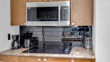 A kitchen with a stainless steel microwave, black tiled backsplash, a black induction cooktop, and wooden cabinets.