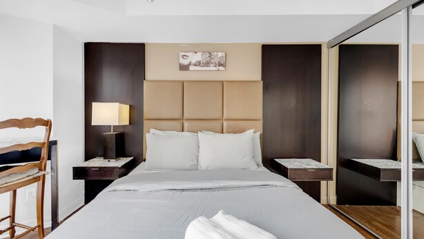 Quarto de hotel com uma cama grande, uma cadeira de madeira, uma mesa de cabeceira com abajur e um espelho.
