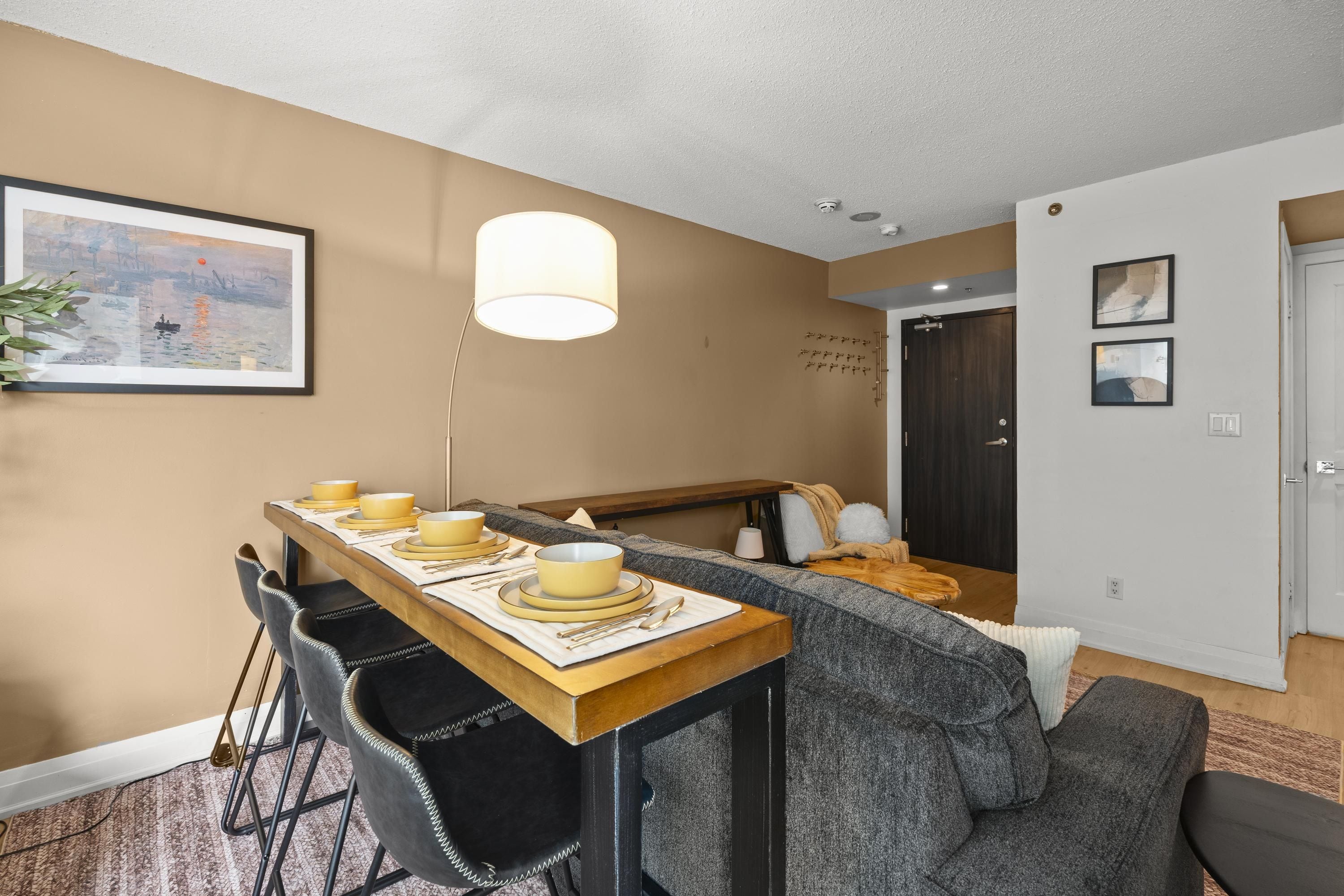 Een eetgedeelte met een houten tafel voor vier personen, een grijze bank en een lijst met een schilderij aan de muur.
