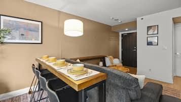 Een eetgedeelte met een houten tafel voor vier personen, een grijze bank en een lijst met een schilderij aan de muur.