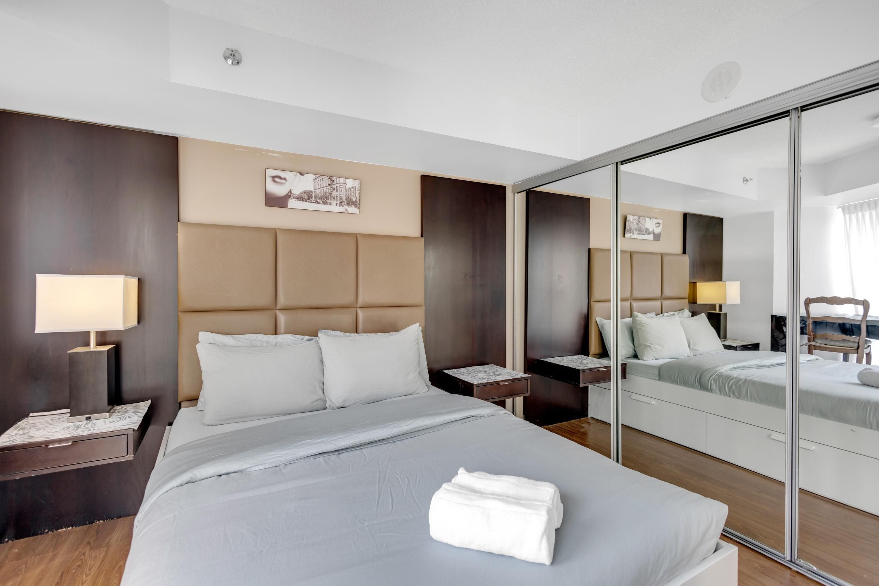A hotel room with a large bed, a bedside table with a lamp, a mirror, and a wooden floor.