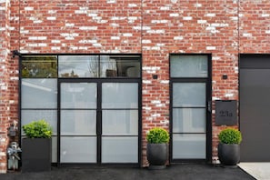 A red brick building with large windows and a garage door. There are three potted plants in front of the windows. - The Pix Studio, Modern Industrial Stay (Greater Geelong)