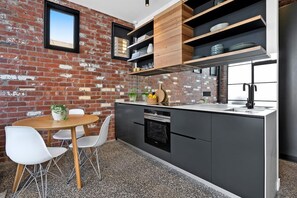 A modern kitchen with a brick wall, wooden shelves, a dining area with white chairs, and a central island with a sink. - The Pix Studio, Modern Industrial Stay (Greater Geelong)