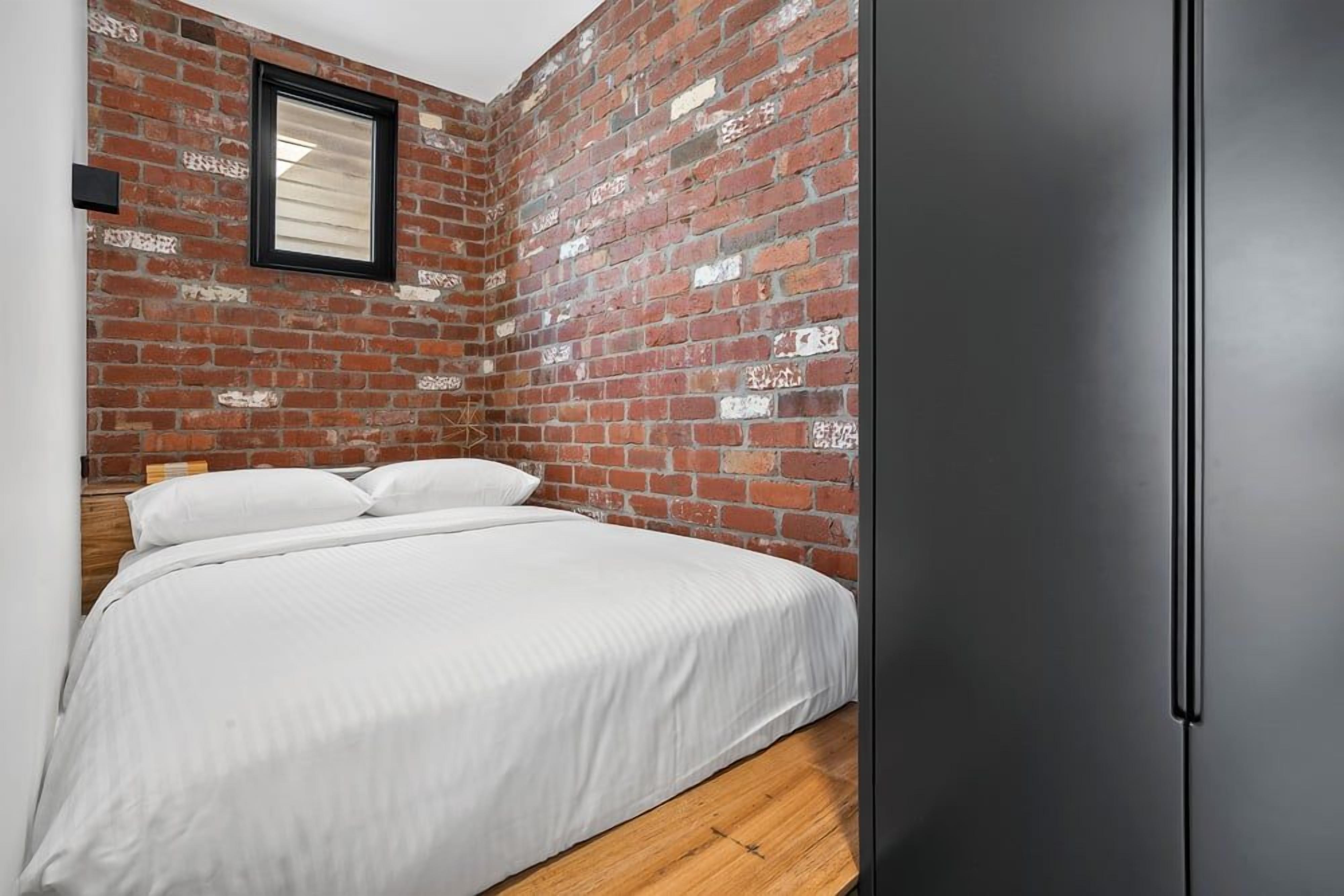 A bedroom with a brick wall, a bed with white bedding, and a black wardrobe.