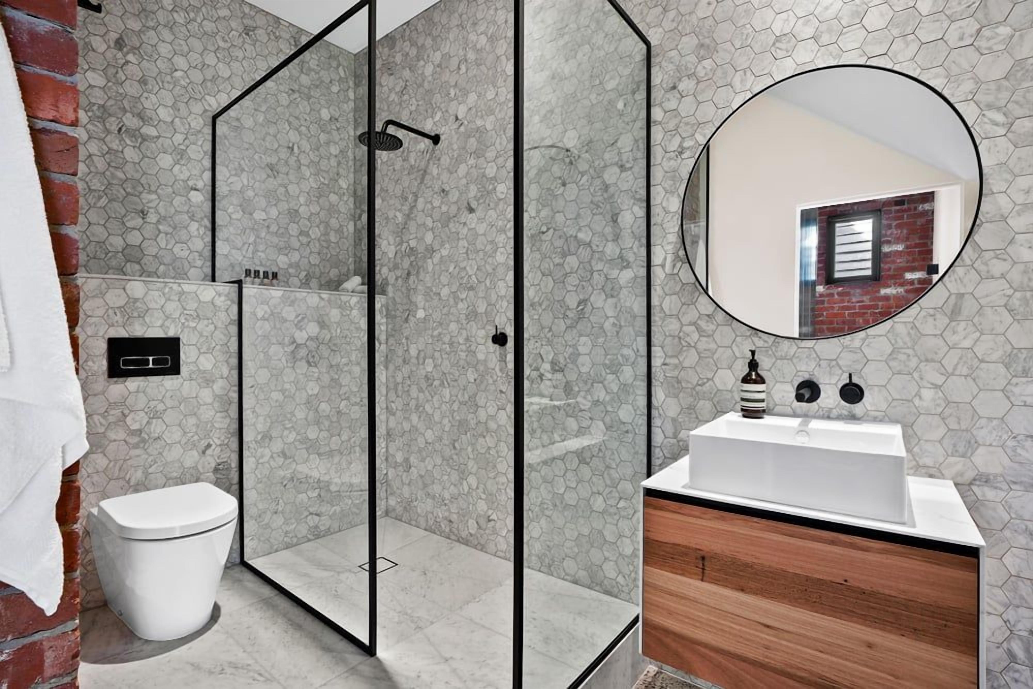 A modern bathroom with a glass-enclosed shower, a white toilet, a wooden vanity, and a round mirror.