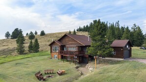 A two-story log cabin with a balcony, surrounded by a grassy field and trees. - Log Cabin Retreat | Private Dock & Mountain Views (Anaconda)