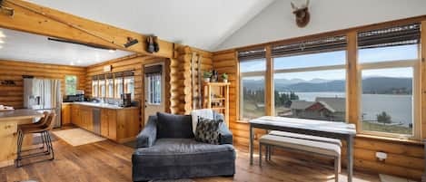 A log cabin interior with a kitchen, dining area, and a large window offering a view of mountains and a lake.
