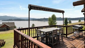 A deck with a table and chairs overlooking a lake, with a view of a cabin and mountains in the distance. - Log Cabin Retreat | Private Dock & Mountain Views (Anaconda)
