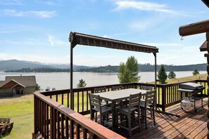 Una terraza con mesa y sillas que da a un lago, con vista a una cabaña y montañas al fondo.