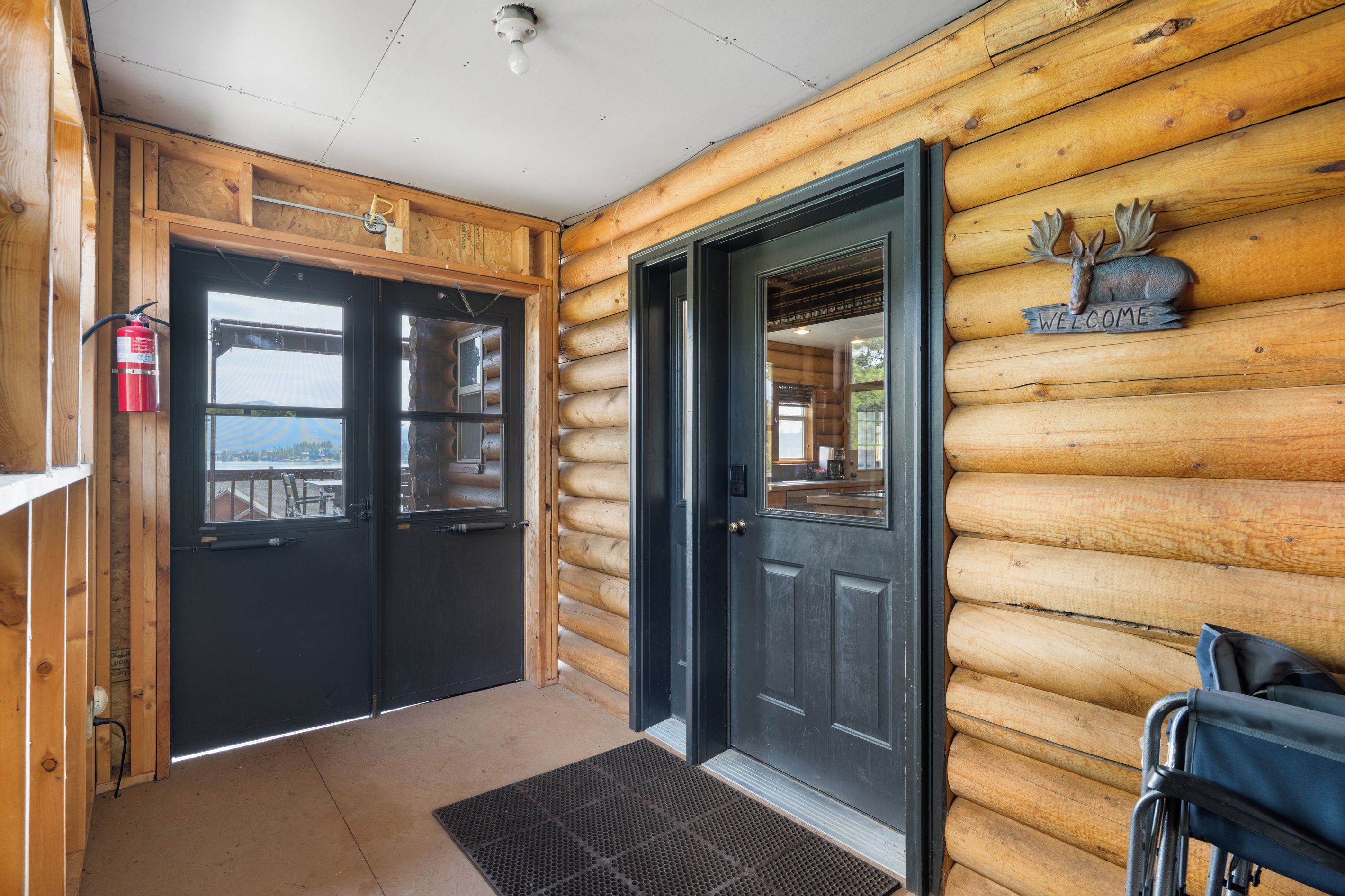 A log cabin with a black door, a fire extinguisher mounted on the wall, and a welcome sign with a moose head.