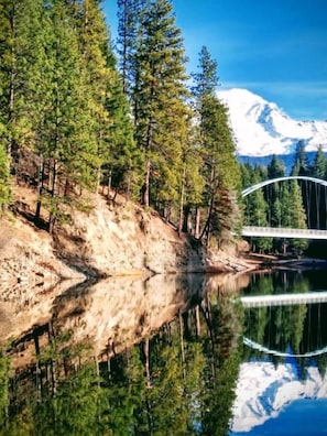 Un lago sereno que refleja árboles y un puente, rodeado por un bosque y una montaña nevada al fondo.