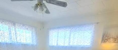 A bedroom with a bed, a nightstand, a lamp, a ceiling fan, and a window with lace curtains.