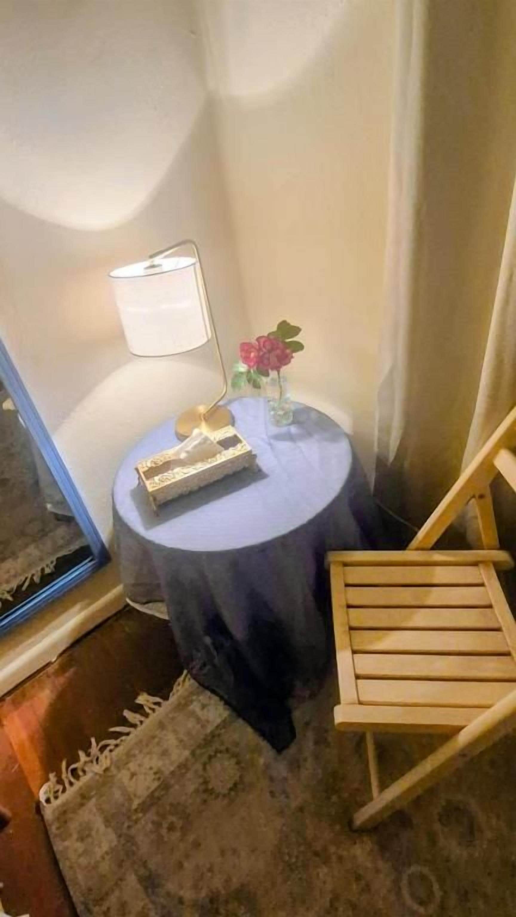 Ett hörnrum med ett litet runt bord, en stol, en lampa och en vas med blommor.