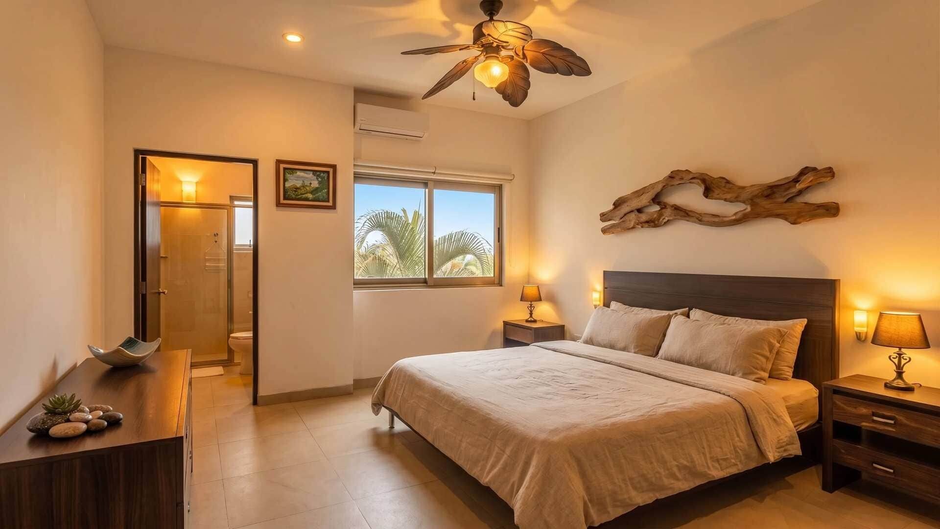 A bedroom with a bed, bedside tables, a ceiling fan, a window with a view, and a door leading to a bathroom.