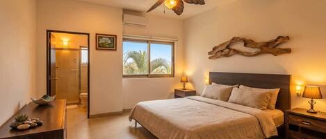 Quarto com cama, mesas de cabeceira, ventilador no teto, janela com vista e porta para um banheiro.