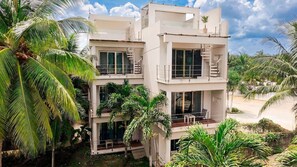 A modern multi-story building with balconies and large windows, surrounded by lush greenery and palm trees. - Maia Ocean Suite at The Beach Club Condos (Quintana Roo)