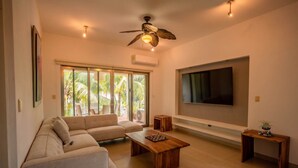 A modern living room with a sectional sofa, a coffee table, a TV mounted on the wall, and a ceiling fan. - Hummingbird Palace at The Beach Club Condos (Quintana Roo)