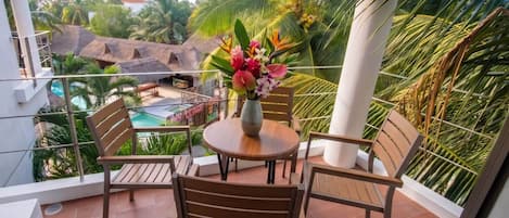 A balcony with a wooden table and chairs, a vase of flowers, and a view of a pool and palm trees.