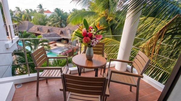 A balcony with a wooden table and chairs, a vase of flowers, and a view of a pool and palm trees.