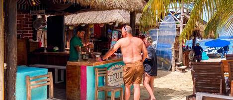 A beachside bar with a thatched roof, wooden furniture, and a sign welcoming visitors.