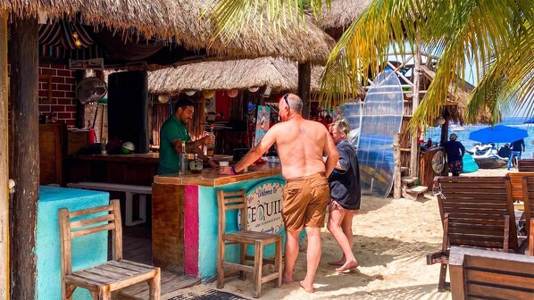 A beachside bar with a thatched roof, wooden furniture, and a sign welcoming visitors.