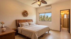 A bedroom with a bed, a bedside table, a decorative wall piece, a window with a view of palm trees, and a door leading to a bathroom.