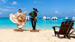 A beach with clear turquoise water, a wooden chair, and a person in traditional attire.