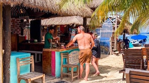 A beachside bar with a thatched roof, wooden furniture, and a sign welcoming visitors.