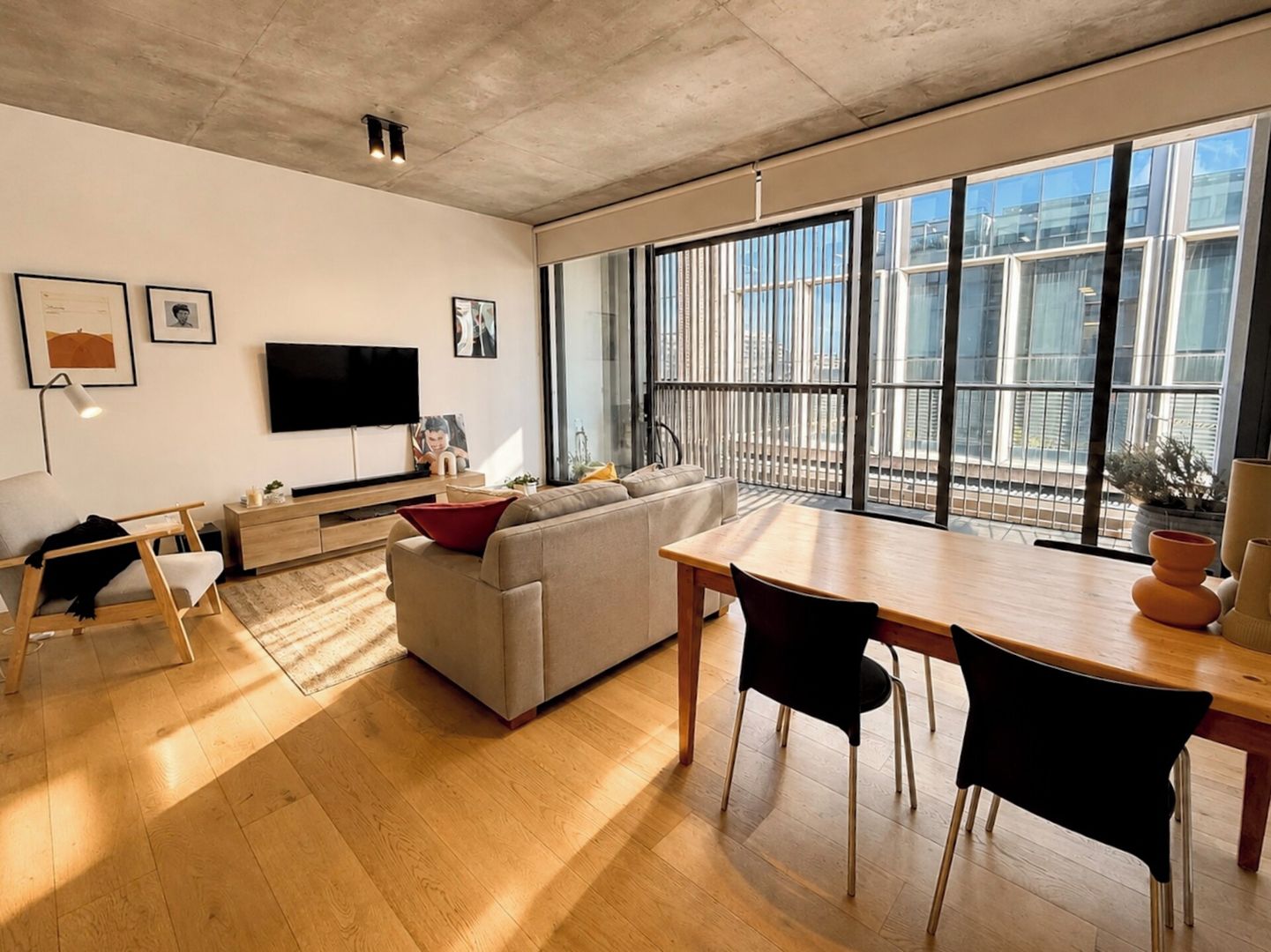 Un salon moderne avec un canapé, une table à manger et des chaises. Un téléviseur est fixé au mur et une fenêtre offre une vue sur les bâtiments.