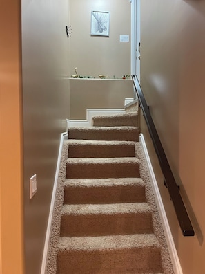 A carpeted staircase with a handrail, a wall-mounted light switch, and a framed picture above the stairs. - Private Luxury Code-Access Suite with Foosball Table (Fort McMurray)