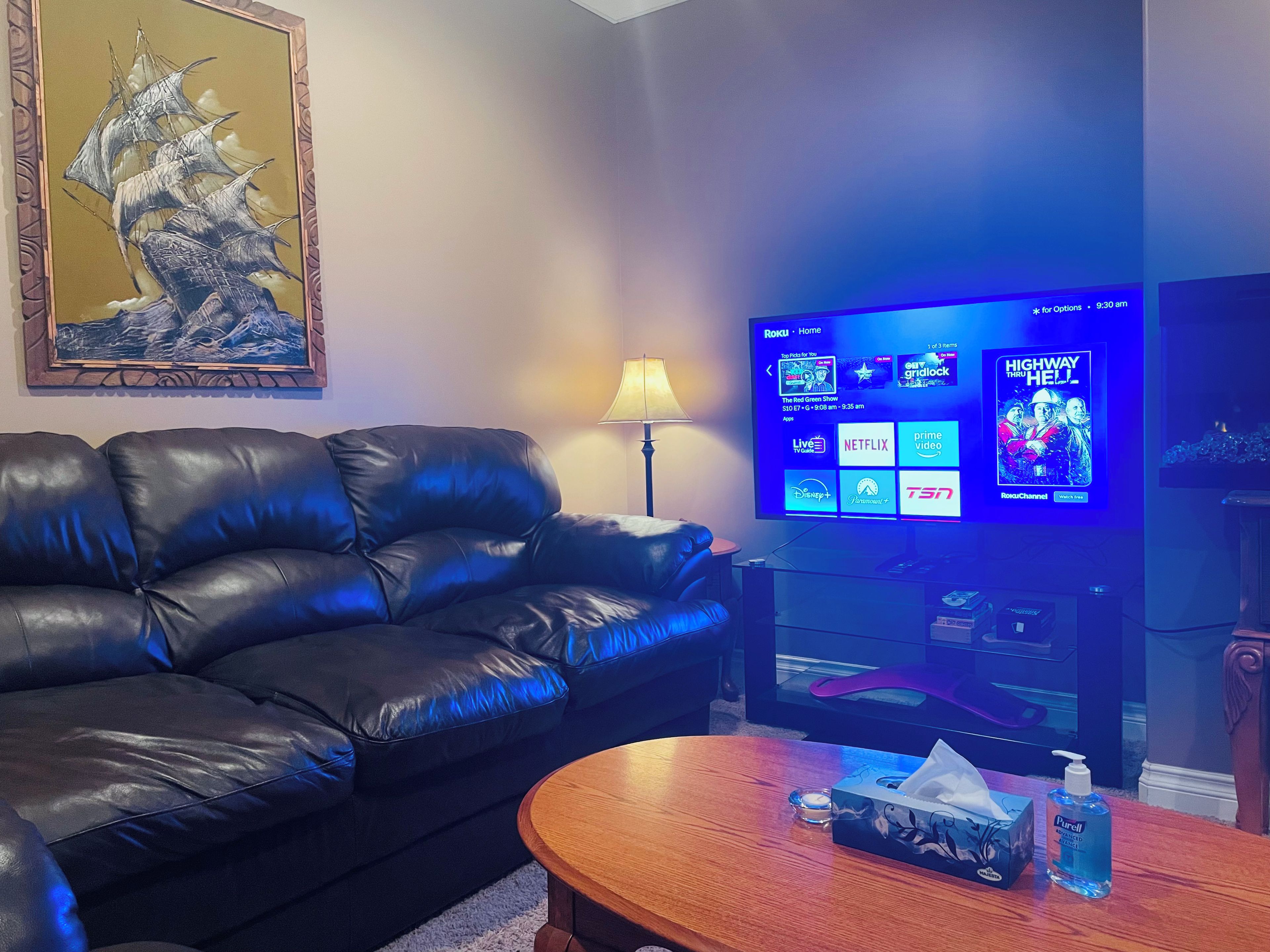 A living room with a leather sofa, a wooden coffee table, a television displaying streaming services, and a framed artwork on the wall.