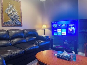 A living room with a leather sofa, a wooden coffee table, a television displaying streaming services, and a framed artwork on the wall. - Private Code-Access Suite with Foosball Table (Fort McMurray)