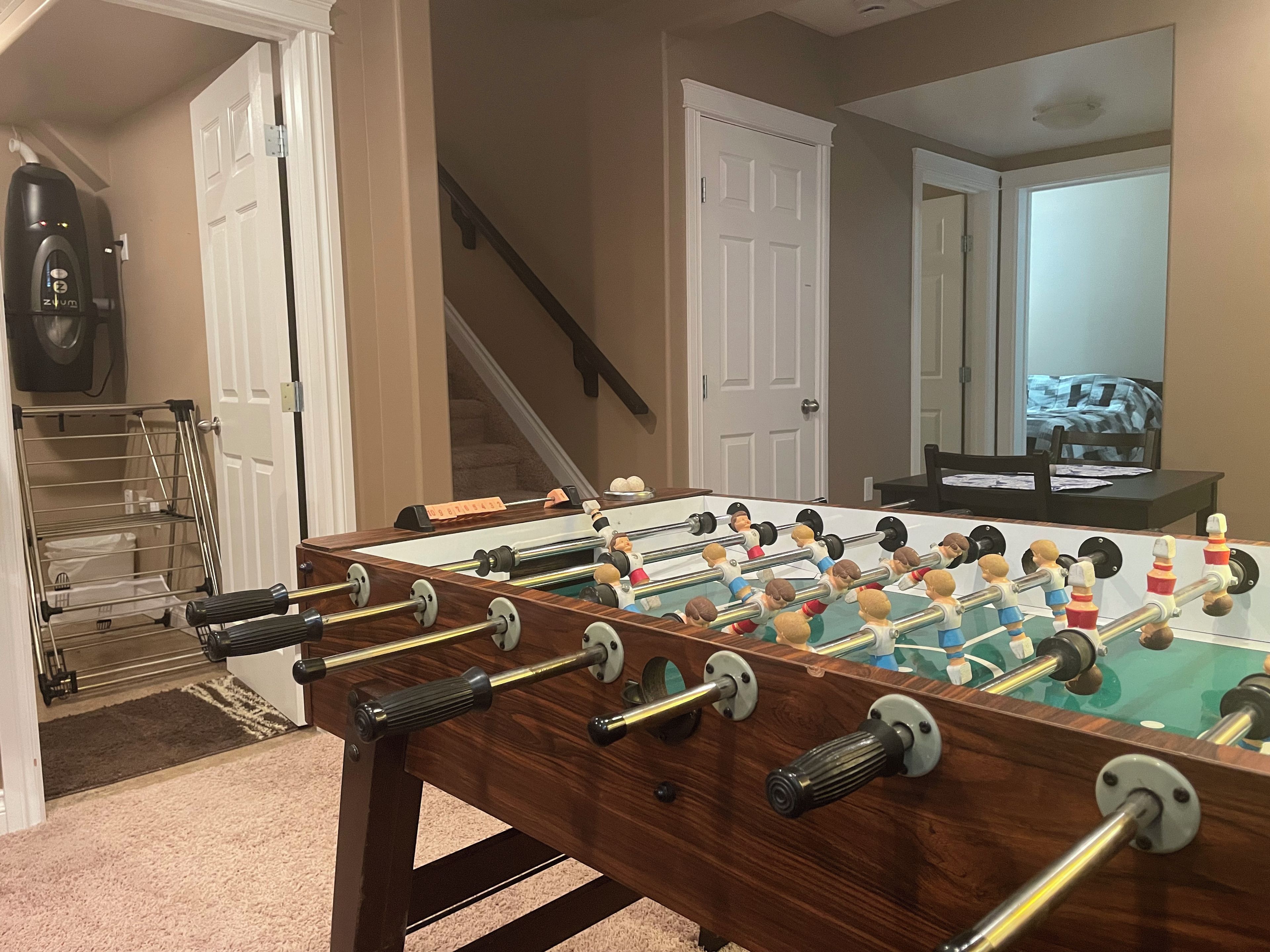 A foosball table in a room with a staircase, a door, and a mirror reflecting a bed.