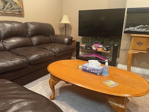 Una sala de estar con un sofá de cuero marrón, una mesa de centro de madera con una caja de pañuelos y una botella, un televisor de pantalla plana y un aparador de vidrio con varios objetos.
