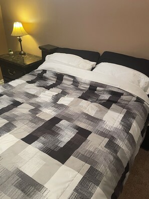 A bed with a checkered quilt, a bedside lamp, and a nightstand with a small plant. - Private Code-Access Suite with Foosball Table (Fort McMurray)
