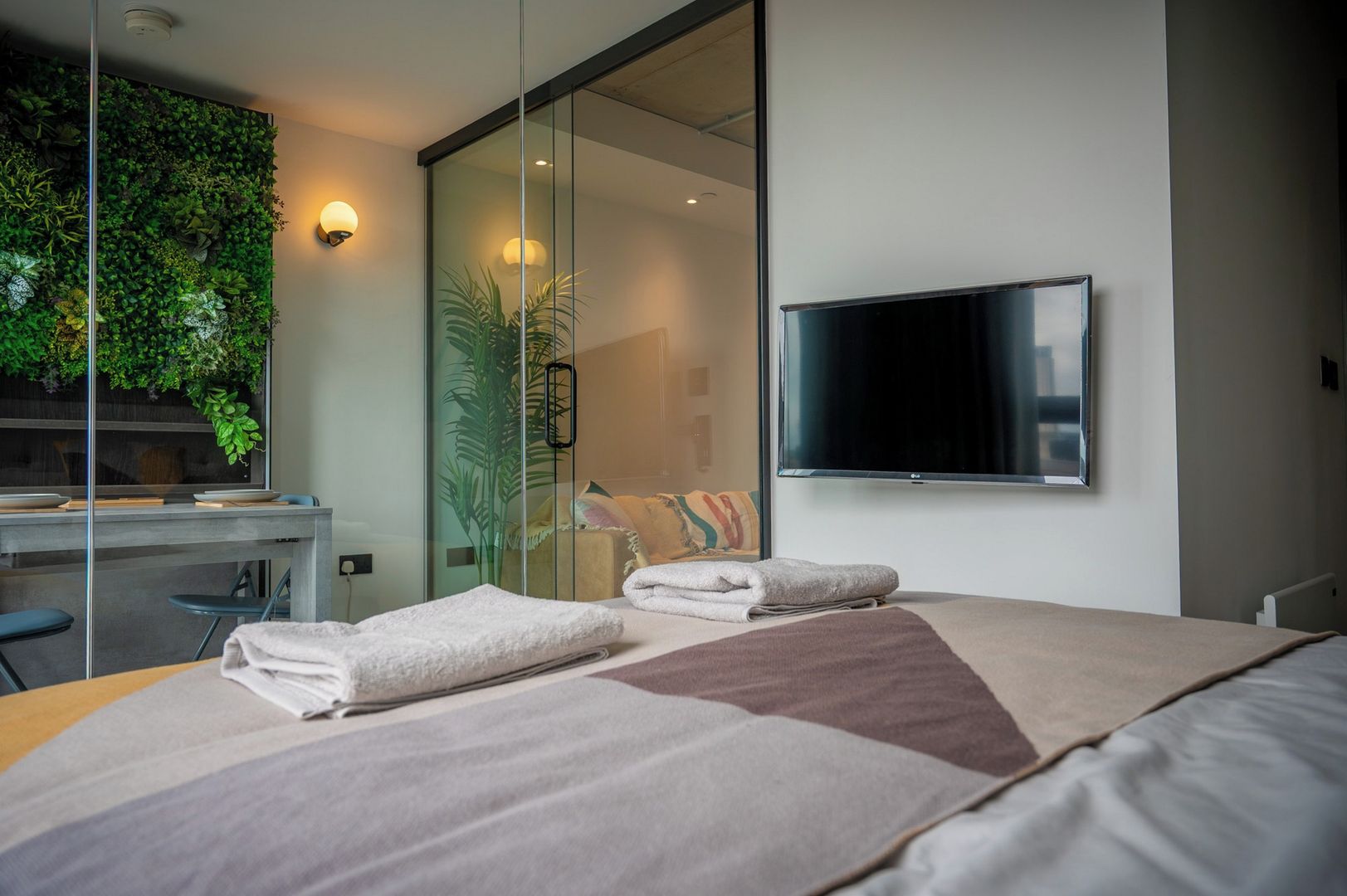 Ett modernt hotellrum med en säng, ett skrivbord, en platt-TV, en glasvägg med en vägg av gröna växter och en skjutdörr.