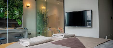 A modern hotel room with a bed, a desk, a flat-screen TV, a glass wall with a green plant wall, and a sliding door.