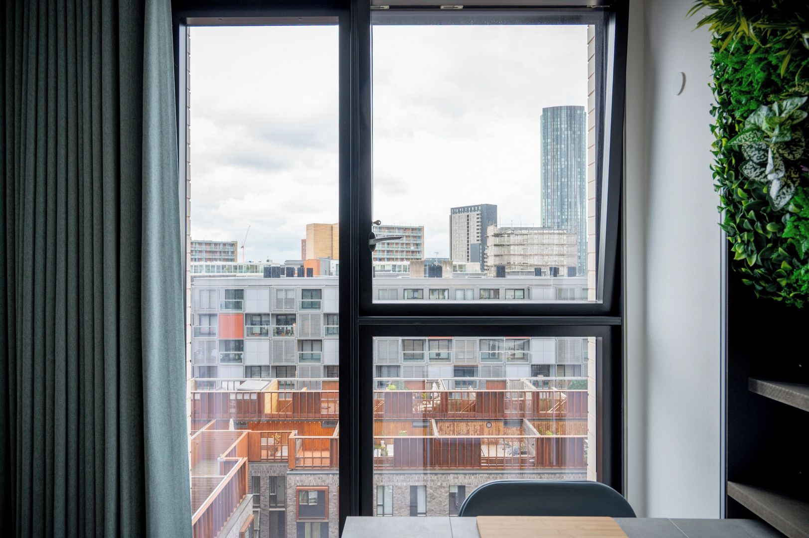 Et værelse med et stort vindue, udsigt til bybilledet, en plante til højre og et bord med stole.