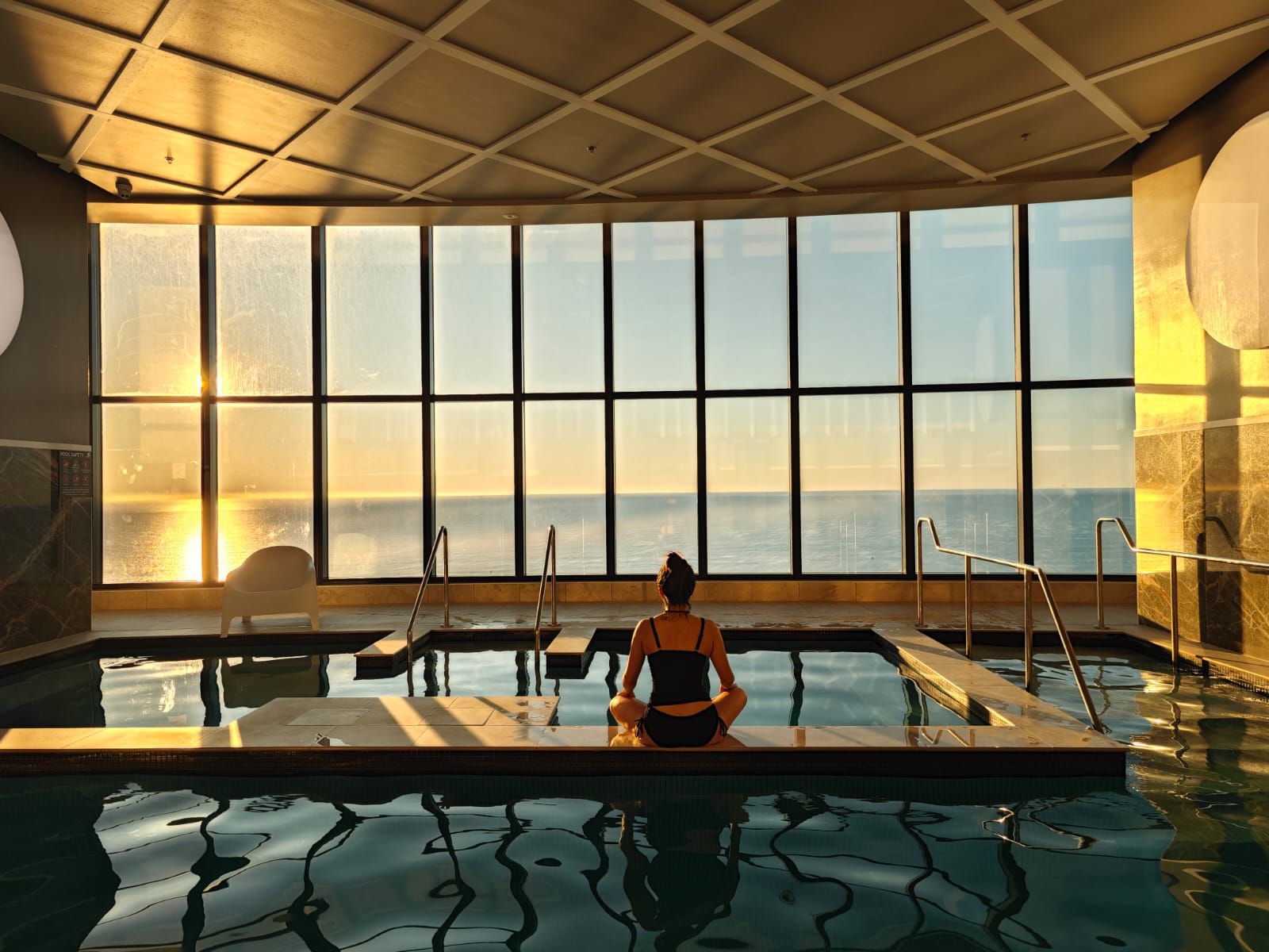 A person meditating by a pool with large windows overlooking the sea.