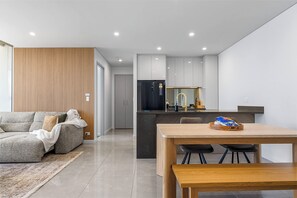 A modern living room with a sofa, a dining table with chairs, and a kitchen area with a refrigerator and sink. - Beachfront Broadwater & Ocean Views 2BR w/ Parking (Surfers Paradise)