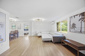A spacious living area with a large white sectional sofa, a dining table with chairs, and a bench. There is a large window providing a view of the outdoors.