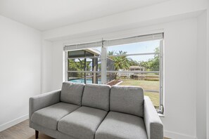 A grey sofa in a room with a large window, white walls, and a view of a swimming pool and palm trees. - Waterfront 2BR · Heated Saltwater Pool & Spa · Dock + 10K Boat Lift (Port Charlotte)