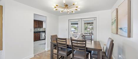 Uma área de refeições com mesa e cadeiras, um lustre moderno e vista através de uma porta de vidro.