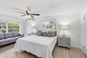 A bedroom with a bed, a nightstand, a lamp, a ceiling fan, and a window with a view of greenery.