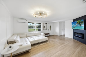 A spacious living room with a white sectional sofa, a small side table, a flat-screen TV mounted on a dark wall, and a modern chandelier.
